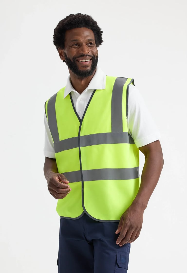 Man smiling wearing a bright yellow-green safety vest with reflective stripes over a white shirt and dark pants